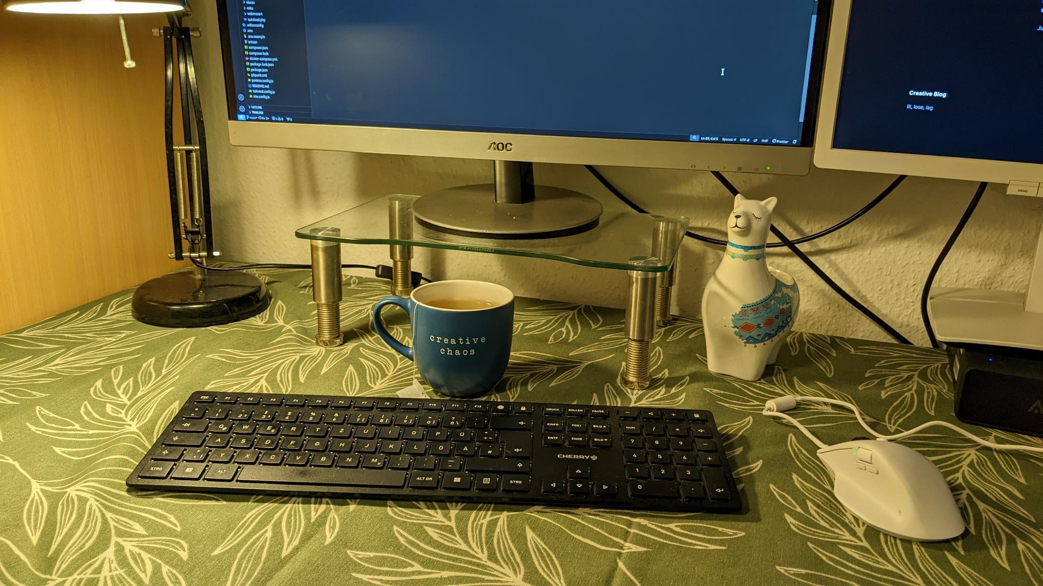 Image of desk with keyboard, mouse, monitors, a cup and imprint "creative chaos" and a cute llama sculpture.
